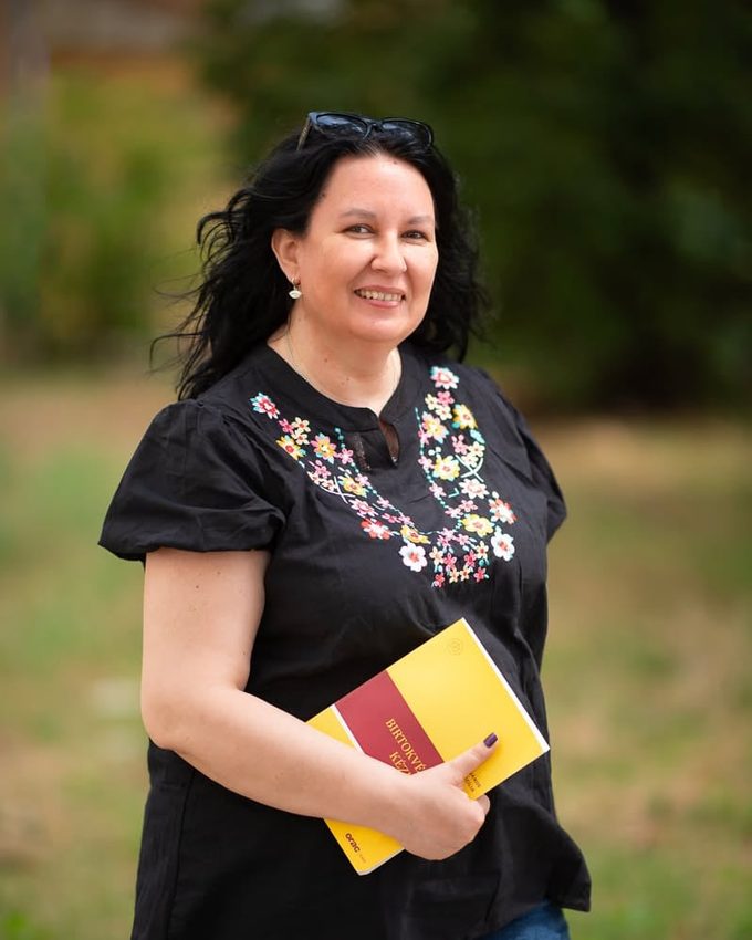 dr. Kajó Cecília mosolyogva áll a szabadban, fekete, színes virághímzéses blúzban; a kezében a sárga borítójú Birtokvédelmi Kézikönyv.