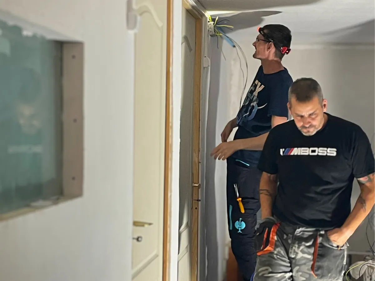 Két férfi villanyt szerel egy félkész, csempézetlen helyiségben: hátul Miki a falon futó vezetékeket nézi, elöl Soma áll